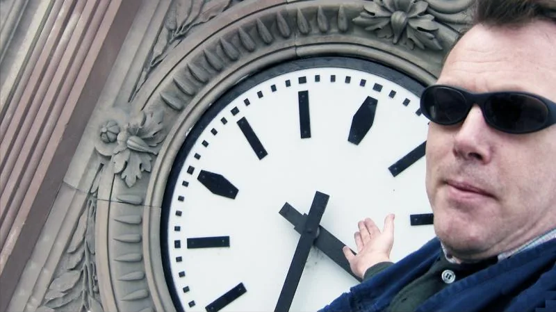 A man in sunglasses points to the hands of a large station clock