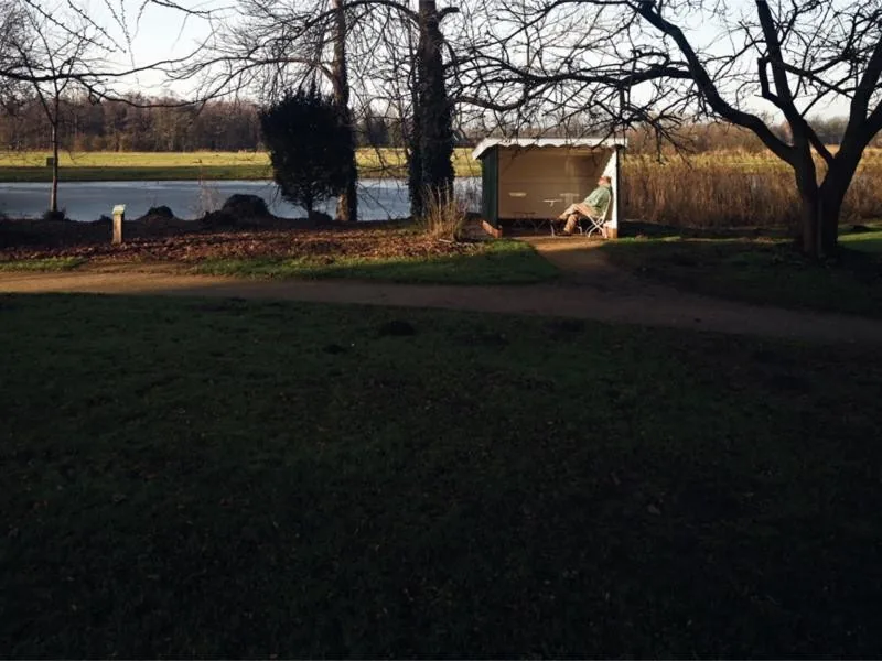 A man sitting in a small shelter beside a frozen pond, under bare trees