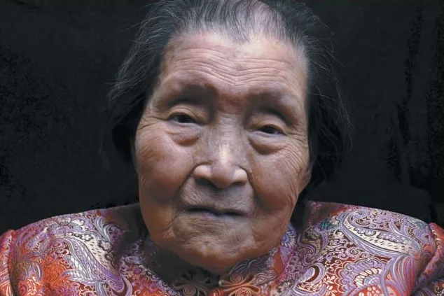 Portrait of an elderly woman in a patterned silk jacket against a dark wall