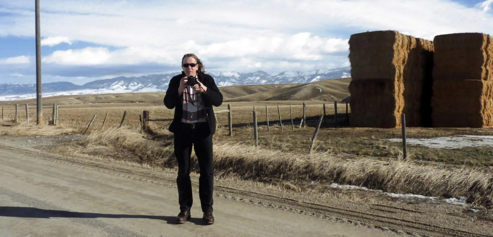 Rob Rombout standing on a gravel road with a camera, hay bales and snowy mountains behind him
